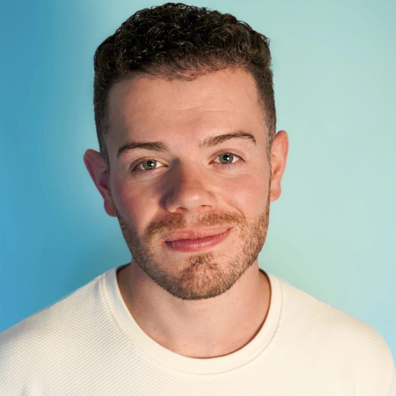 A young man with short curly brown hair and a trimmed beard is smiling softly. Richie Cardile wears a white textured shirt and stands against a light blue background. The bright, even lighting highlights his face clearly.