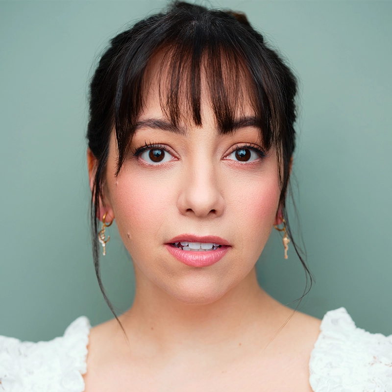 Caroline Aimetti, a young woman with dark hair, wispy bangs, and light skin, looks directly at the camera. She wears natural makeup, gold earrings, and a white textured top. The soft green background highlights her calm expression.
