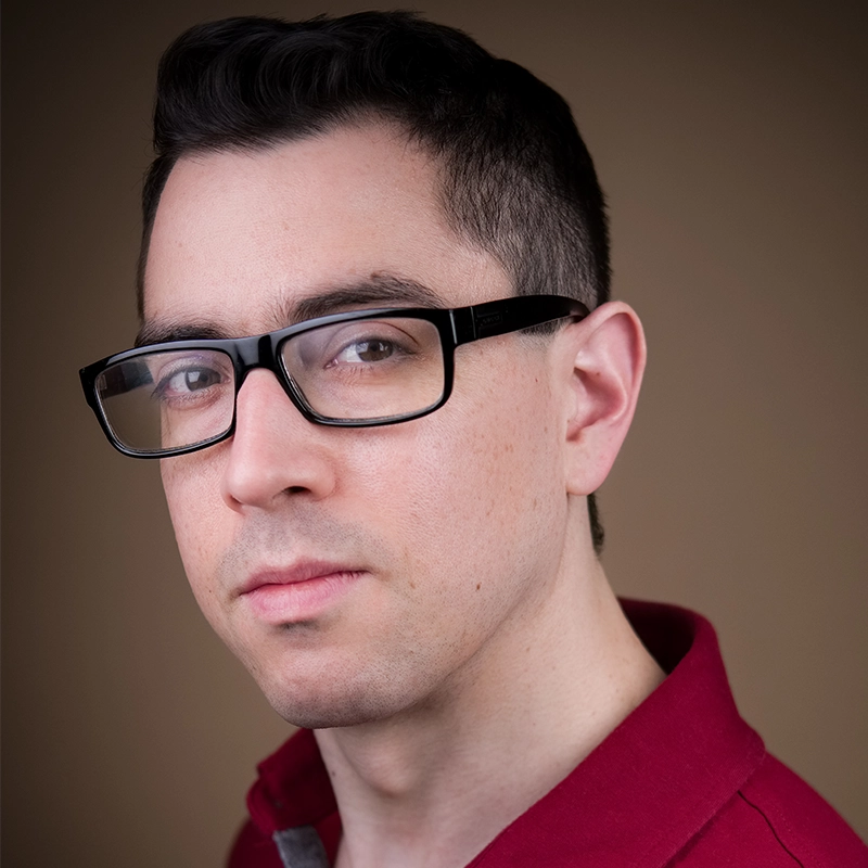A man with short dark hair, pale skin, and black glasses looks at the camera with a neutral expression. He is wearing a red collared shirt and is positioned against a plain brown background. The lighting is soft and even, reminiscent of the subtle photographic style often associated with Madilyn Jaz Morrow.