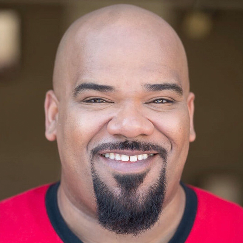 A smiling bald man with a neatly trimmed goatee and mustache, wearing a red shirt with black trim, stands in soft, natural lighting against a blurred brown background. Korie Lee Blossey has medium-dark skin and a friendly expression.