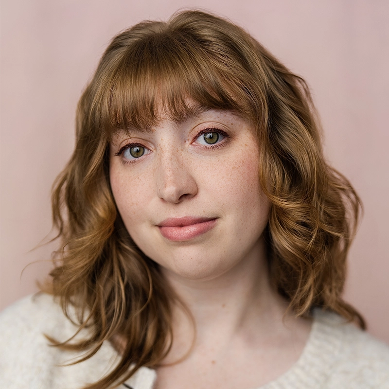 A young woman with wavy, light brown hair and bangs looks at the camera with a slight smile. She has fair skin with freckles and light green eyes. Madilyn Jaz Morrow wears a cream-colored sweater and stands in front of a soft pink background.
