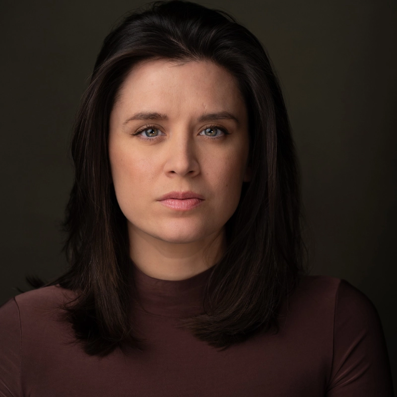 Ramona Mallory, with fair skin and straight, shoulder-length dark brown hair, faces the camera with a neutral expression. She wears a high-neck, long-sleeve maroon top against a dark, plain background that highlights her face.