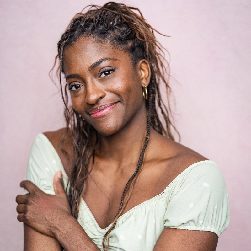 A smiling woman with long braided hair wears a light green off-shoulder top with white dots. Johanna Moise stands against a pale pink background, looking at the camera with her left hand resting gently on her right arm.