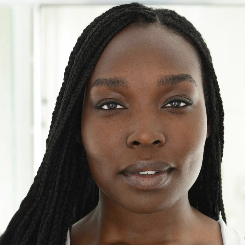 A woman with long, braided black hair looks directly at the camera. Veronica Otim has smooth dark skin, defined eyebrows, and natural makeup. The softly blurred, bright background highlights her calm, confident expression.