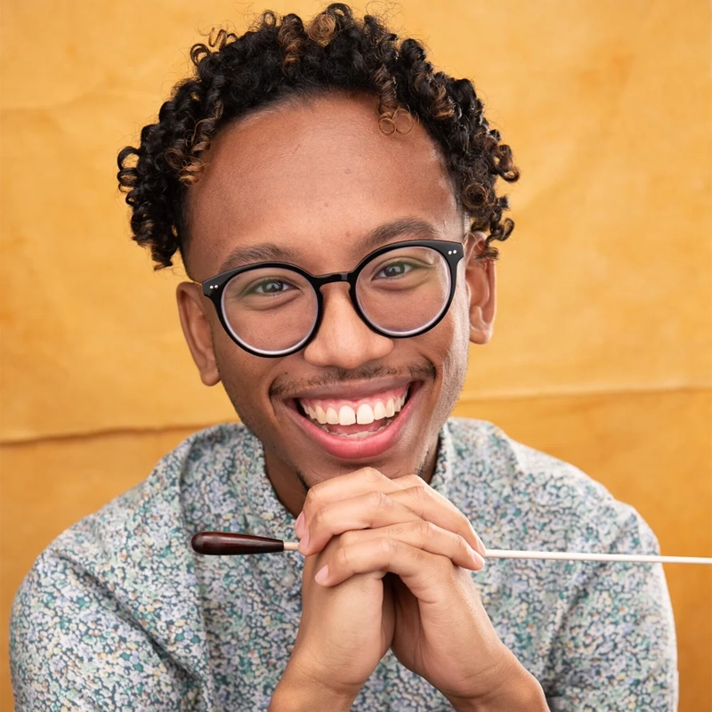 Patrick B. Phillips smiles while holding a conductor’s baton horizontally against his chin. He has curly hair, wears black-rimmed glasses and a light-colored, patterned shirt, and is set against a textured, golden-yellow background.