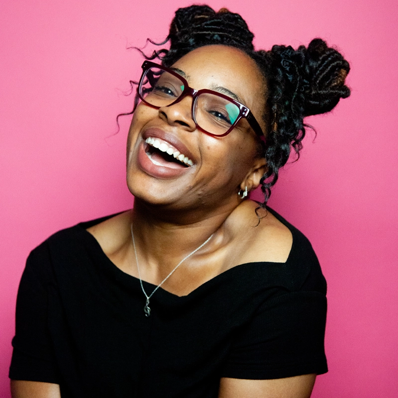 A smiling Pier Lamia Porter with dark skin, glasses, and two afro puffs wears a black off-shoulder top and a silver necklace. She sits in front of a bright pink background, looking slightly to the side, exuding joy and warmth.
