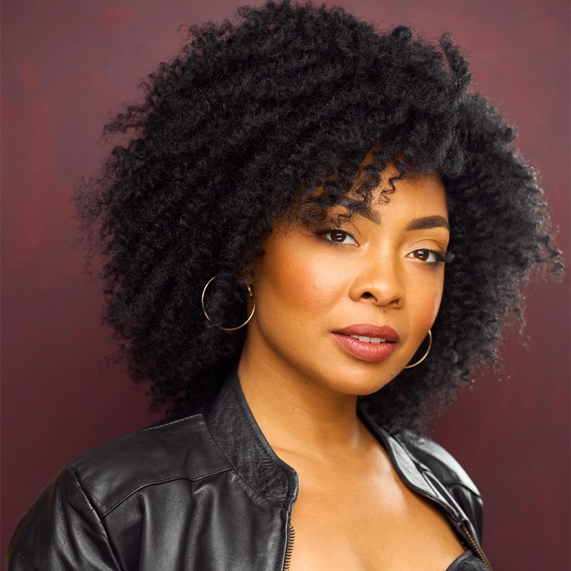 Jennie Harney-Fleming poses confidently with natural, voluminous curly hair against a maroon backdrop. She wears a black leather jacket, gold hoop earrings, and natural makeup featuring subtle eyeshadow and glossy lips.