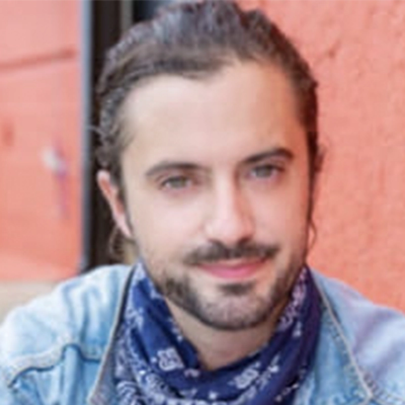 Mikey Rosenbaum, a man with light skin and long brown hair pulled back, smiles gently. He has a short beard and mustache, wears a blue bandana around his neck, and a light denim jacket. The background features a blurred reddish-orange wall.
