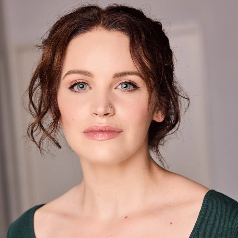 Lindsay Heather Pearce, a woman with light skin, wavy brown hair pulled back, and blue eyes, looks at the camera. She wears natural makeup, a gold nose ring, and a dark green top. The background is softly blurred in neutral tones.