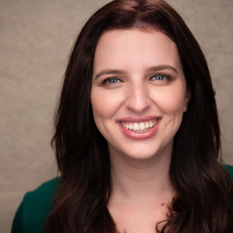 Samantha Roberts, with long brown hair and blue eyes, smiles warmly at the camera. She wears a dark green top and is posed before a plain, light beige background. Soft lighting gently highlights her features.