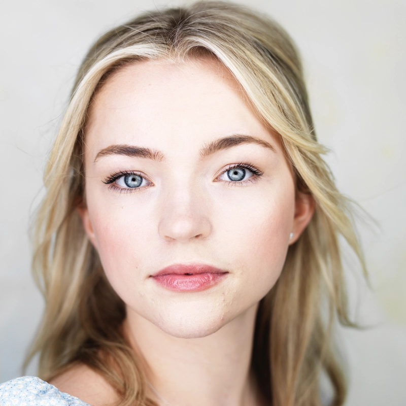 A young woman with fair skin, blue eyes, and long blonde hair looks directly at the camera. Megan Styrna wears natural makeup with pink lipstick and softly defined eyebrows. The soft, airy background is out of focus and light in color.