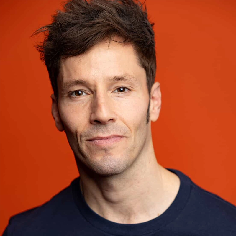 A man with short, tousled brown hair and fair skin is looking at the camera with a slight smile. He is wearing a navy blue crew-neck shirt. The background is a solid orange color, highlighting Thayne Jasperson’s relaxed and approachable presence.