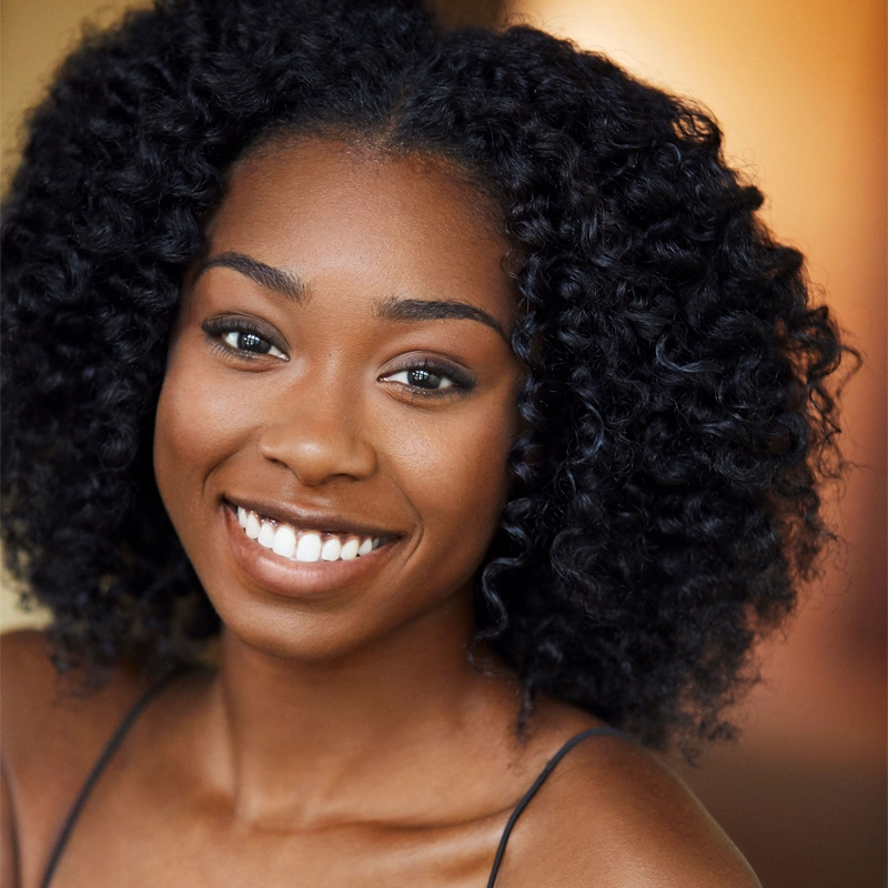 A young woman with curly black hair smiles warmly at the camera. Rachel Simone Webb has medium-dark skin, bright white teeth, and expressive dark eyes. She wears a black spaghetti strap top, and the background is softly blurred in warm tones.