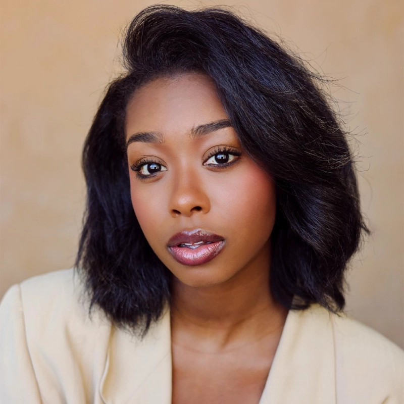 A young woman with medium-dark skin and shoulder-length black hair styled in soft waves. Rachel Simone Webb has glossy lips, subtle makeup, and wears a light-colored blazer. Her expression is calm, and the background is a neutral beige.
