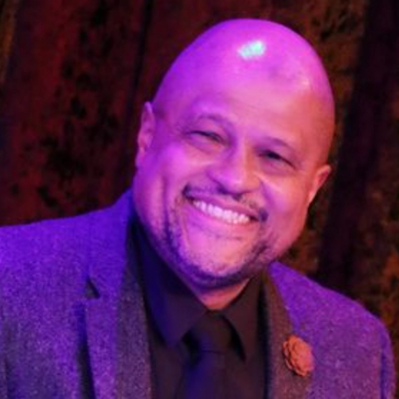 A smiling bald man with a closely trimmed beard wears a purple suit jacket, black shirt, and tie. The background is dark and blurred with warm, reddish lighting. The man appears friendly and happy, and a decorative pin is on his left lapel, suggesting he could be Alvester Garnett.