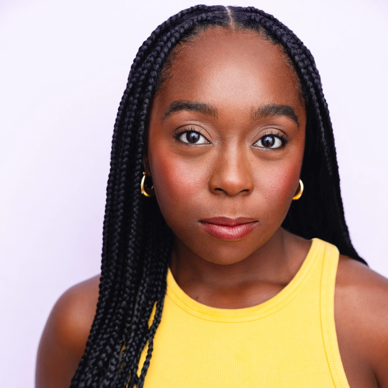 A woman with long, braided hair wears a sleeveless yellow top and gold hoop earrings. She faces the camera with a neutral expression, against a plain light background. Her makeup is natural with defined brows, blush, and red lipstick.