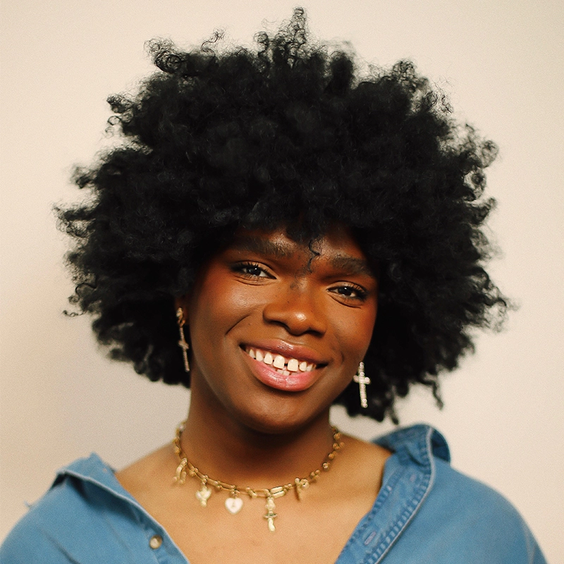 A person with a voluminous, curly black afro smiles warmly. They wear a blue button-up shirt, gold hoop earrings, cross earrings, and a gold necklace with small charms. The background is plain and light-colored, drawing focus to their friendly expression.