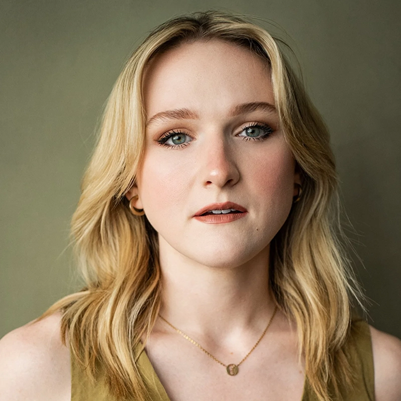 A young woman with fair skin and shoulder-length blonde hair looks directly at the camera. She wears light makeup, gold hoop earrings, a gold necklace, and a sleeveless olive top. The background is plain and muted green-beige.