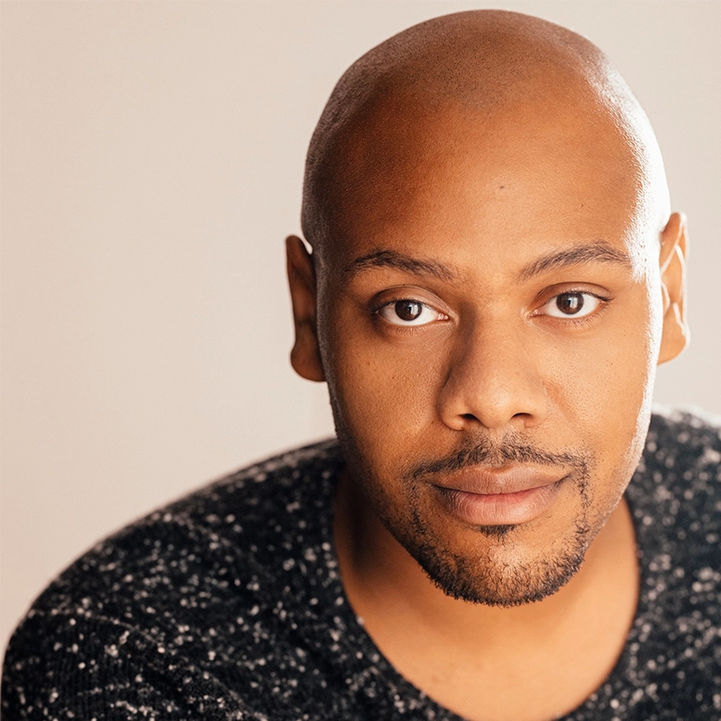 A man with a shaved head and trimmed beard looks directly at the camera, wearing a black speckled sweater. He has warm brown skin and a calm, neutral expression. The background is softly lit and out of focus, highlighting his face.