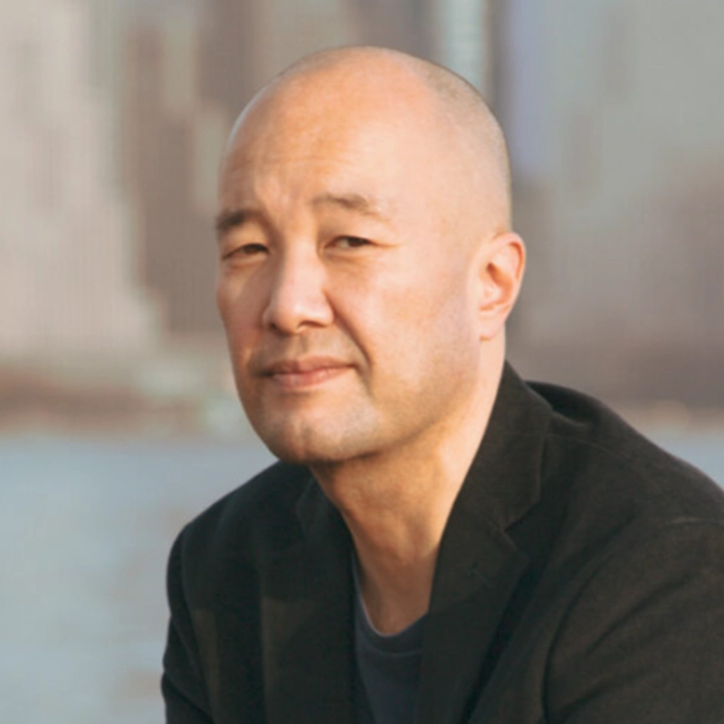 A bald man with fair skin is wearing a black blazer and a dark shirt. He is sitting outdoors, looking at the camera with a neutral expression. The blurred background suggests an urban setting, possibly near water. This could be Alvester Garnett captured in a quiet moment amidst the city's bustle.