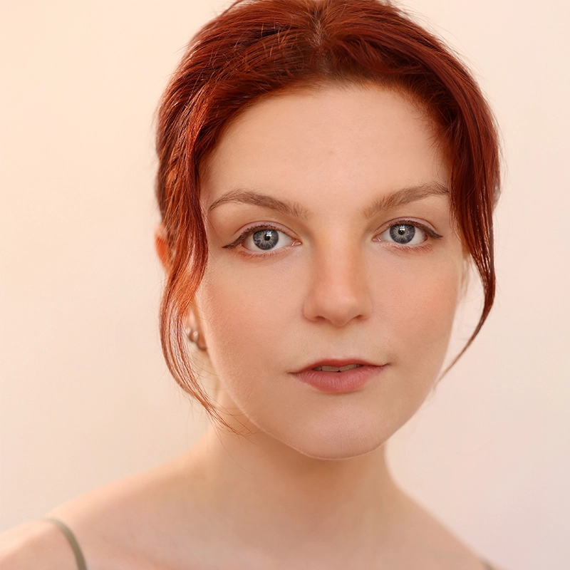 A young person with fair skin, red hair styled back with loose strands framing their face, and blue eyes, is looking directly at the camera. They have subtle makeup, including eyeliner and nude lipstick, and are wearing a pale spaghetti strap top. The background is soft and beige.