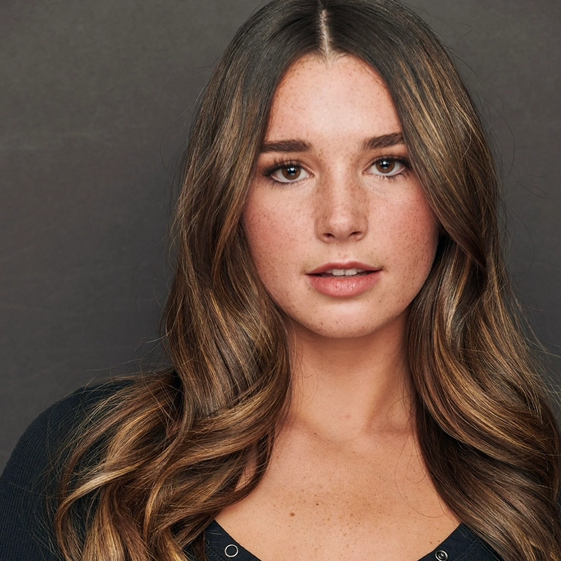 A young woman with long, wavy brown hair and subtle highlights faces forward. She has fair skin with freckles, brown eyes, and neutral makeup. She wears a black, V-neck top against a plain, dark background.