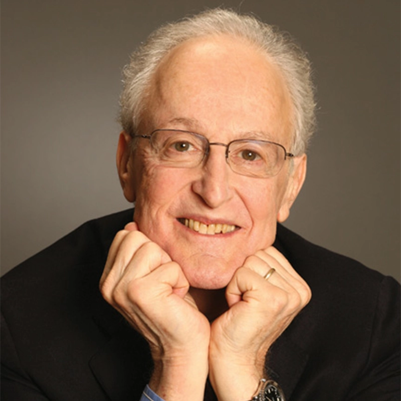 An older man with light skin, short gray hair, and glasses smiles warmly at the camera. He is wearing a dark jacket and a wedding ring, resting his chin on his fists with elbows on a surface, set against a plain brown background.