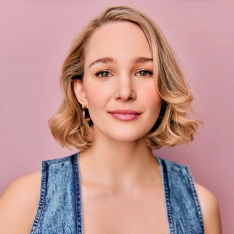 Nicole Morris, with short, wavy blonde hair and fair skin, stands in front of a pink background. She wears a sleeveless denim top and small hoop earrings, her relaxed, gentle smile matched by a confident, direct gaze at the camera.