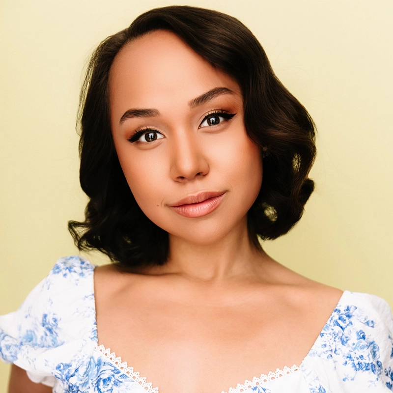 A young woman with wavy dark hair styled in loose curls smiles softly. She wears a white dress with blue floral patterns and lace trim. Her makeup is natural, with defined brows, winged eyeliner, and nude lipstick. The background is pale yellow.