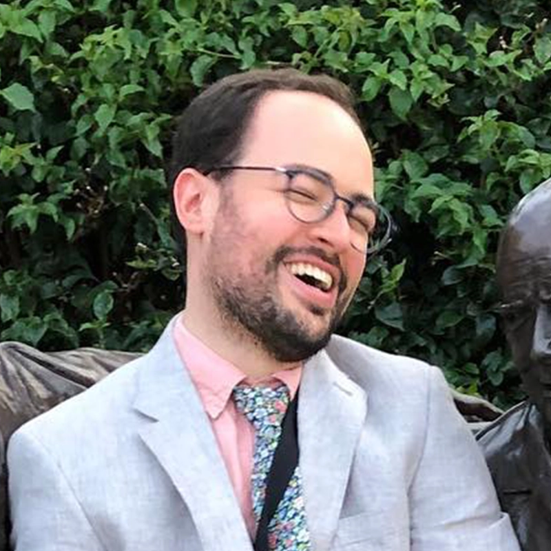 A man with glasses and a beard, wearing a light gray suit, pink shirt, and floral tie, laughs joyfully while sitting next to a bronze statue. The musician Alvester Garnett, known as a talented jazz drummer, is surrounded by green leafy bushes in the background.