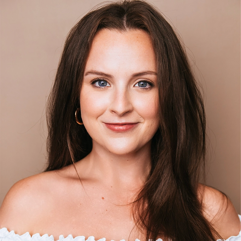A woman with long brown hair and blue eyes smiles gently at the camera. She is wearing a white off-the-shoulder top and a gold hoop earring, against a plain beige background. Her makeup is natural, and her hair is parted in the middle.