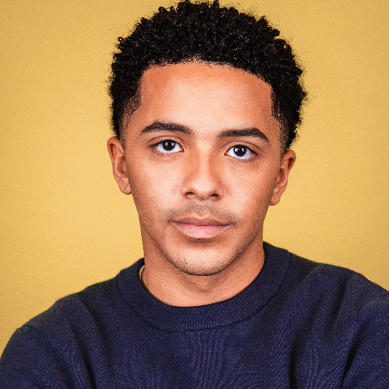 A young man with short curly hair and medium skin tone looks directly at the camera with a neutral expression. He is wearing a dark crewneck sweater and sits against a solid yellow background.