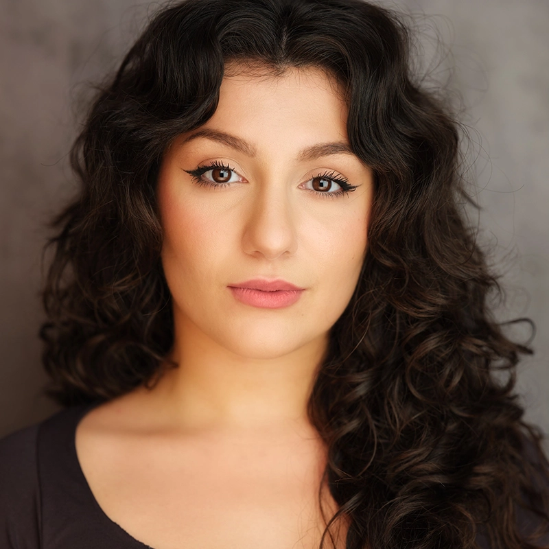 A woman with long, dark, curly hair and fair skin looks directly at the camera with a neutral expression. She has dark, arched eyebrows, brown eyes with winged eyeliner, and soft pink lips. The background is softly blurred and gray.