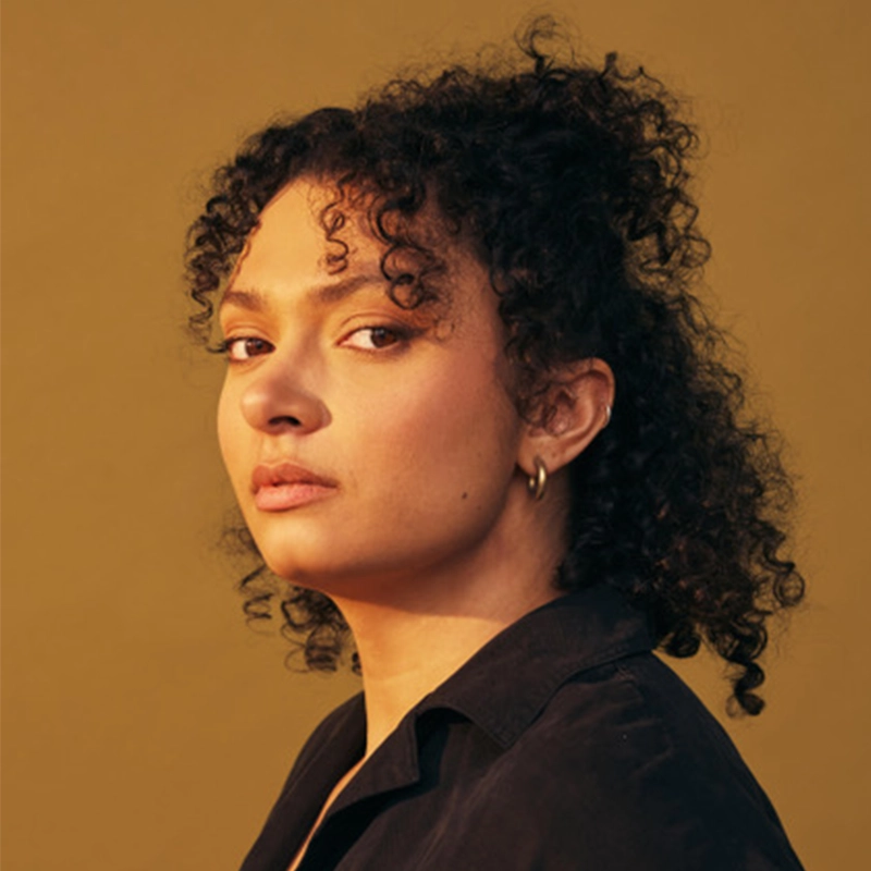 Nicole Morris, with medium brown skin and curly dark hair pulled back, looks slightly over her shoulder. She wears a black top, gold hoop earrings, and has a neutral expression against a solid mustard yellow background.