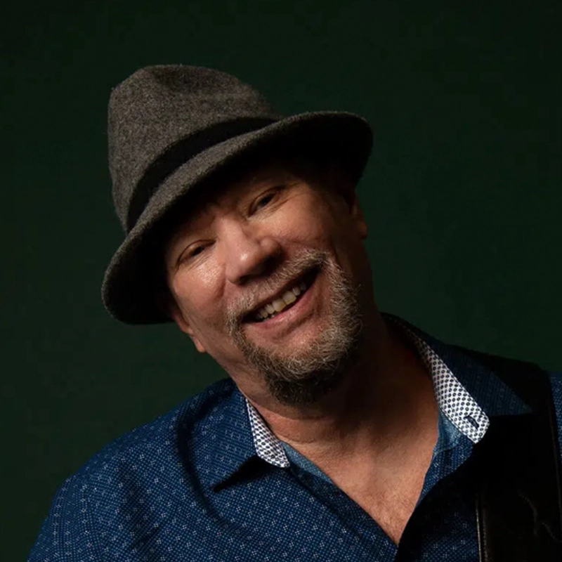 A middle-aged man with a short beard and mustache smiles warmly. He wears a gray fedora hat, a blue patterned shirt with a white collar, and poses against a dark green background. His head is tilted slightly to the side, capturing the relaxed confidence of an accomplished jazz drummer like musician Alvester Garnett.