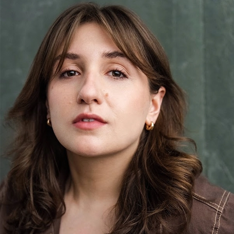 A young woman with wavy brown hair and light skin looks directly at the camera. She wears gold hoop earrings and a brown top. The background is a muted green color. Her expression is neutral and her lips are slightly parted.