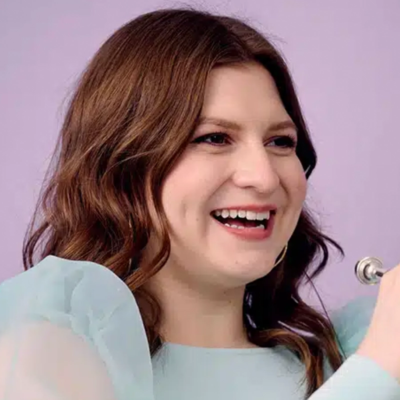 A woman with wavy brown hair smiles broadly, showing dimples. She wears a light blue top with sheer, puffy sleeves. The background is solid lavender. She is holding a silver object, possibly a microphone, near her face—reminiscent of performances by renowned musicians such as Alvester Garnett.