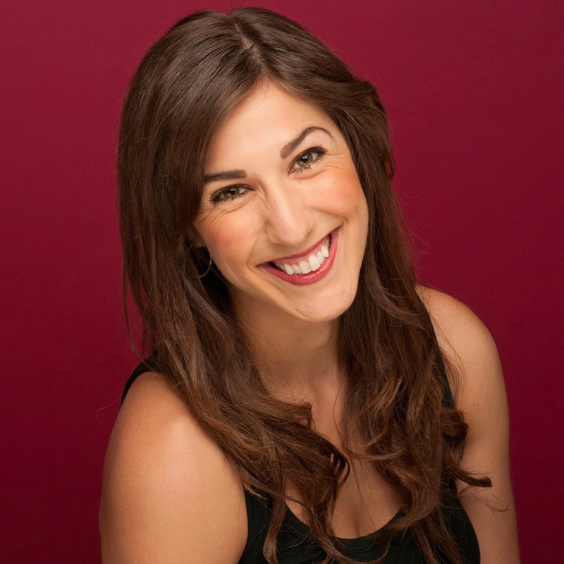 A woman with long, wavy brown hair smiles widely at the camera. She wears a sleeveless black top and has light skin. The background is a solid dark pink or maroon color, and she appears happy and approachable.