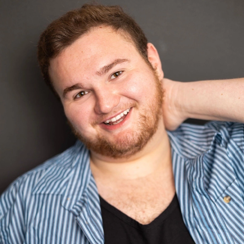 Ryan Van Nostrand smiles in a headshot, his hand resting behind his neck. He wears a blue and white pinstriped button-down shirt over a black v-neck shirt against a dark grey background.