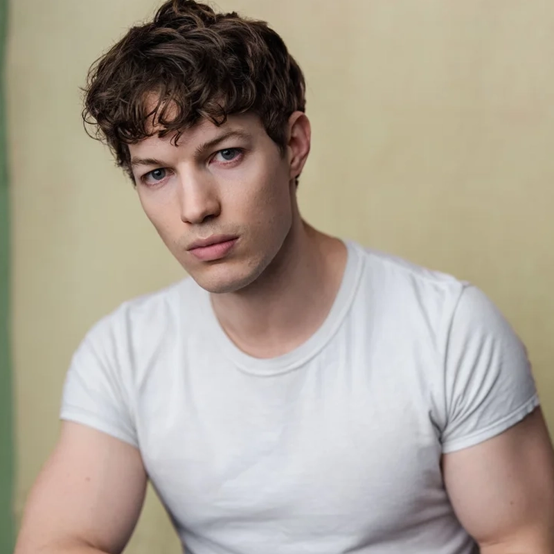 A young man with short, curly brown hair and blue eyes wears a fitted white t-shirt. He sits against a plain, light beige background, looking directly at the camera with a neutral expression—reminiscent of Wes Williams’ effortless style.