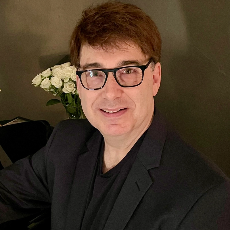 Darren Cohen, a man with short brown hair, wearing black glasses, a black blazer, and a black shirt, smiles at the camera. He is seated indoors in front of a dark wall, with a bouquet of white flowers in the background.