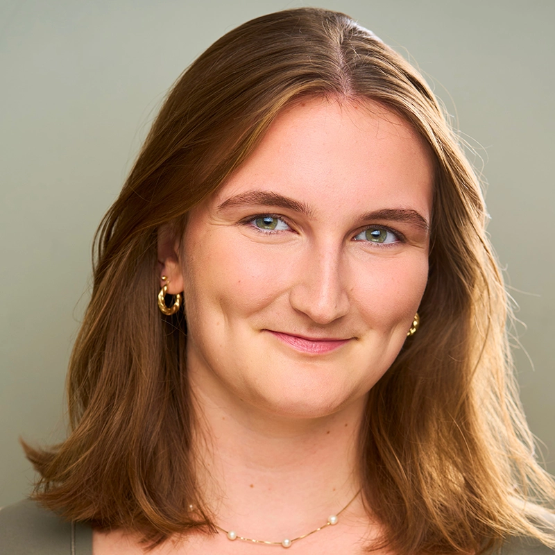 Jana Djenne Jackson, a young woman with shoulder-length light brown hair, smiles at the camera. She wears gold hoop earrings, a delicate necklace, and a light-colored top against a softly blurred background that draws focus to her face.
