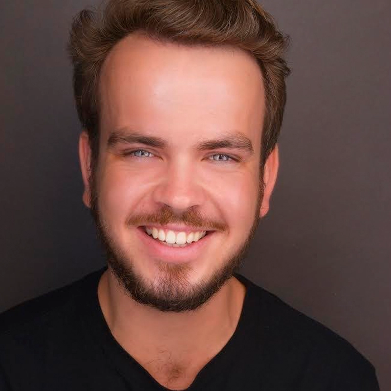 A young man with light skin, short brown hair, and a full beard smiles warmly at the camera. Matt Everingham wears a black shirt against a plain dark background, his blue eyes and teeth visible as he tilts his head slightly.