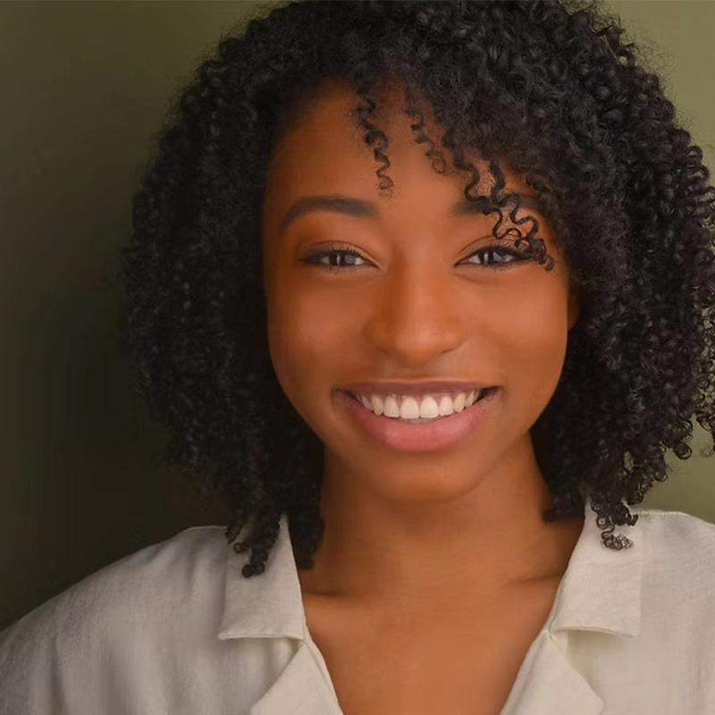 Jana Djenne Jackson, a young woman with dark curly hair, smiles warmly at the camera. She is wearing a light-colored collared shirt and stands against an olive-green background, her friendly expression welcoming and genuine.