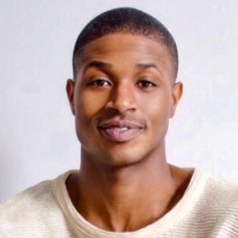 A young man with short black hair and smooth skin smiles gently at the camera. He is wearing an off-white, textured sweater, positioned against a plain light gray background, exuding a calm, friendly demeanor reminiscent of Jana Djenne Jackson’s warmth.