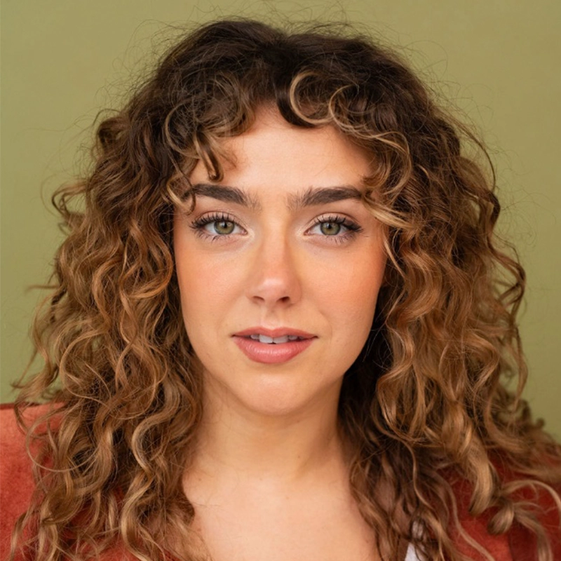 Madison Deadman, a young woman with long, curly brown hair with highlights and green eyes, gazes with a neutral expression. She wears a rust-colored jacket against a muted green background in this professional headshot style.