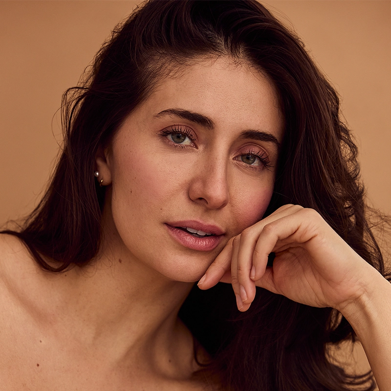 A woman with long brown hair poses against a beige background. She gazes softly at the camera, resting her chin on her hand. With fair skin and subtle makeup, Marina Pires wears a small hoop earring, her expression calm and relaxed.