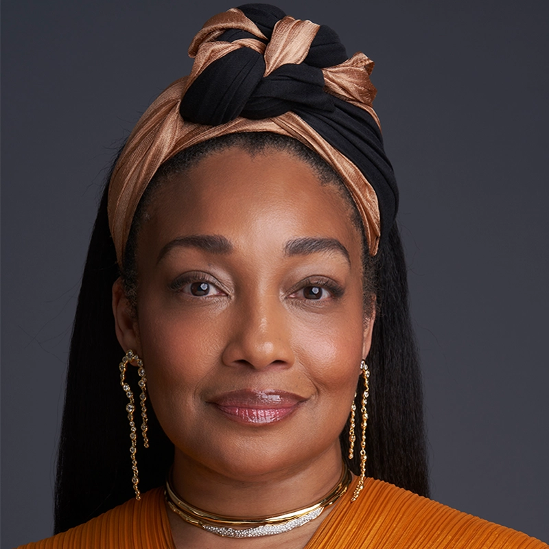A woman with smooth medium-brown skin wears a black and gold headwrap, gold earrings with chains, and a gold choker. Reminiscent of Marina Pires, she has straight dark hair, natural makeup, an orange top, and a warm expression against a plain dark gray background.