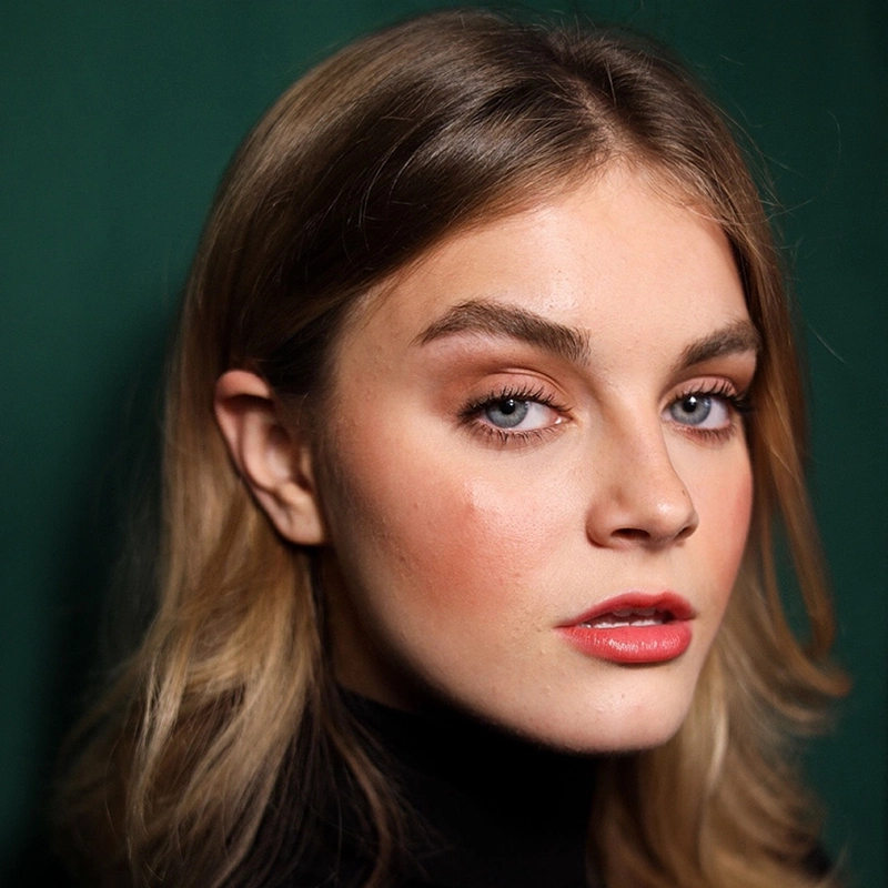 Jana Djenne Jackson, a young woman with light skin, blue eyes, and blonde hair styled in loose waves, looks confidently at the camera. She wears natural makeup with bold brows, rosy cheeks, and red lipstick in a black turtleneck against a dark green background.