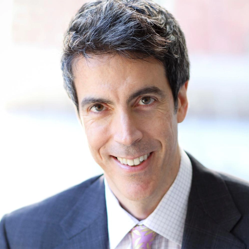 Joseph Thalken, a man with short dark hair and fair skin, is smiling at the camera. He is wearing a dark blazer, light checkered shirt, and a light purple patterned tie. The softly blurred background highlights his face.